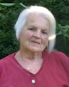 Photo of Denise before her passing. Denise wearing a red shirt sitting in front of green foliage. She has white chin length hair.